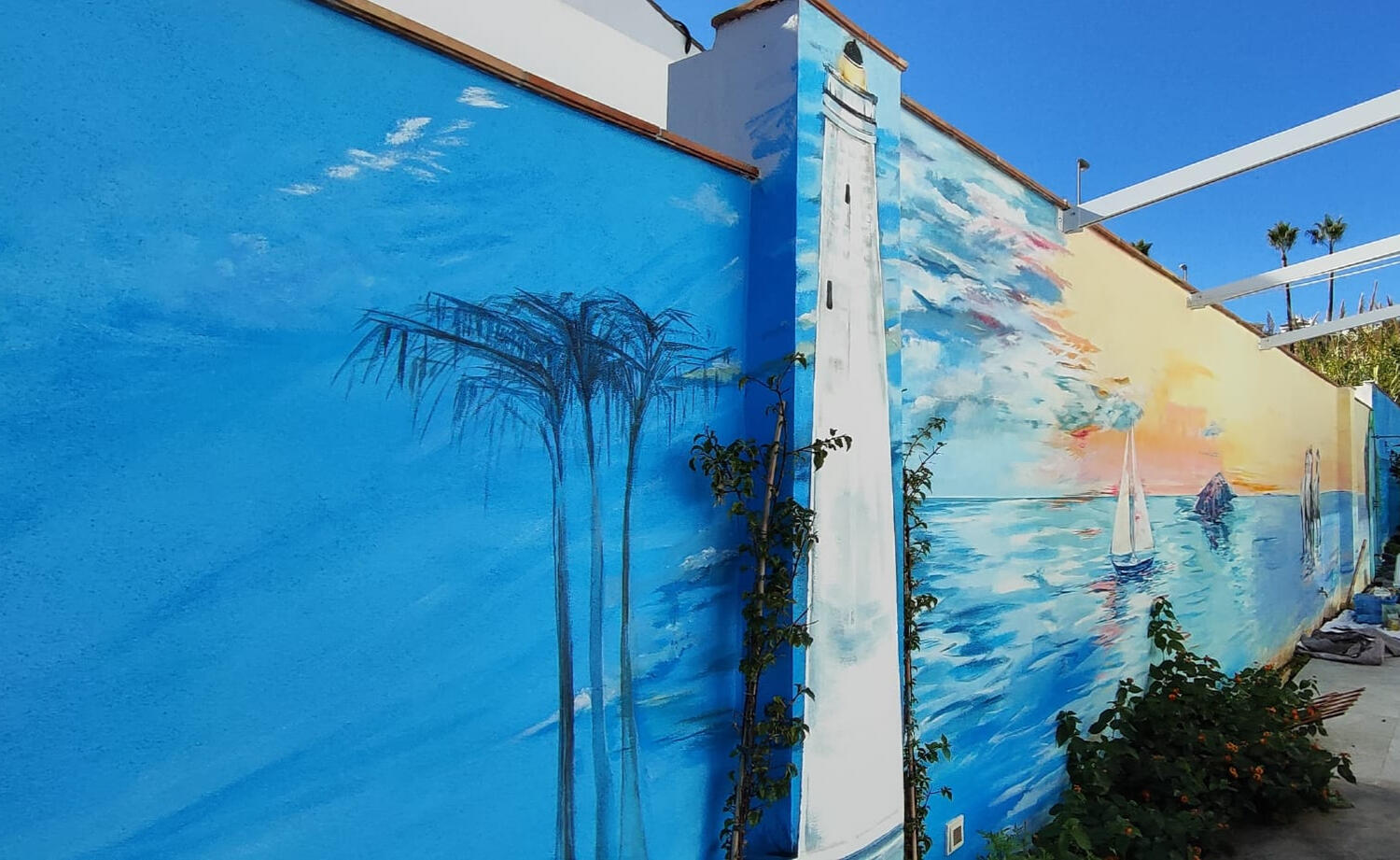 Mural con palmeras y el faro pintado en la columna, En la pared, a su lado, el mural del velero y una pequeña isla en un atardecer naranja y veraniego. Pintado por Lol Malone en La Cala de Mijas para Tooker Vacation Properties.